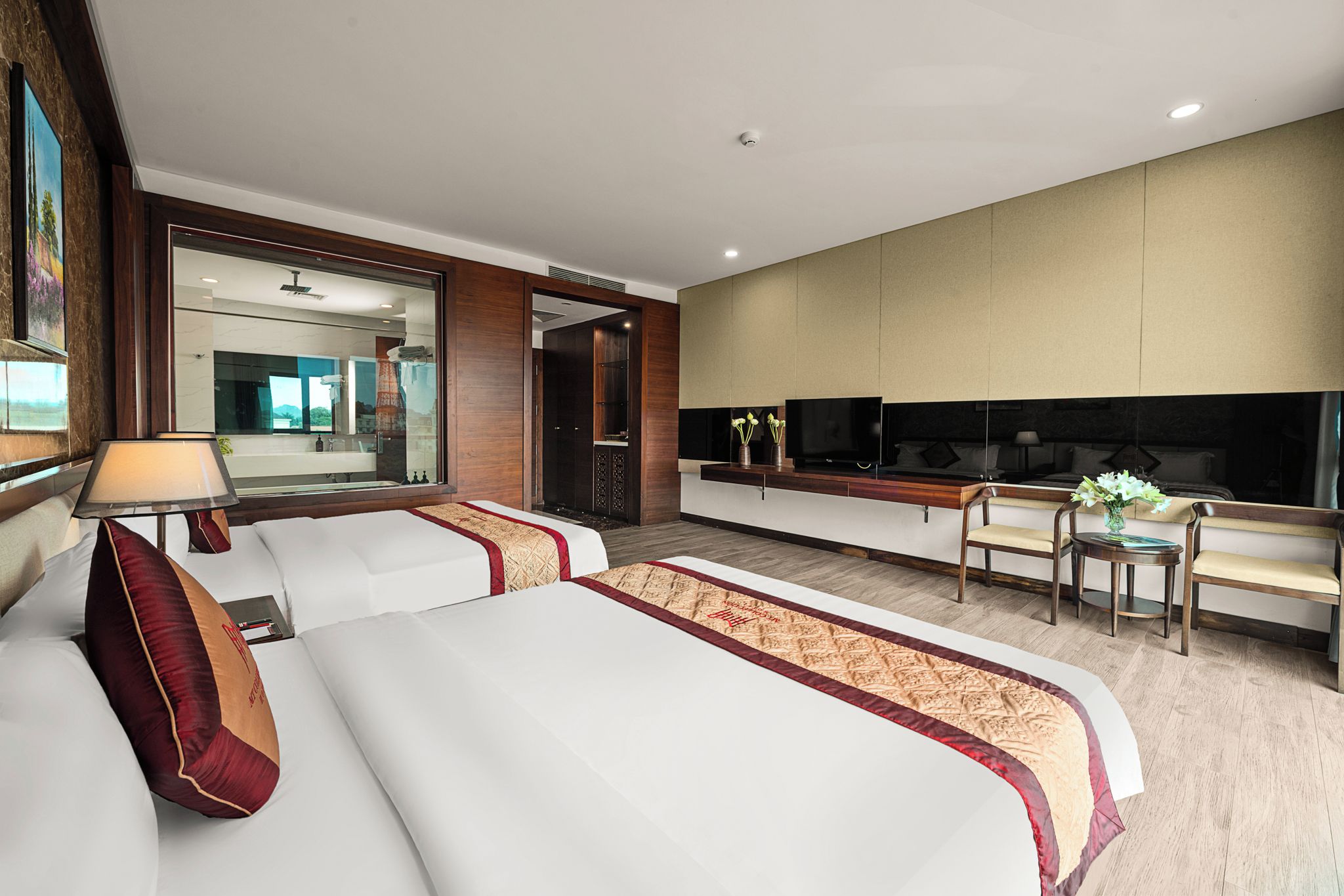 Habitación de hotel con dos camas, una zona de estar con una mesa y sillas, un espejo grande y un televisor.