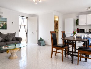 A modern living room with a glass coffee table, a sofa with cushions, a dining area with a wooden table and chairs, and a kitchen with white cabinets and appliances. - Townhouse with shared pool (Torrevieja)