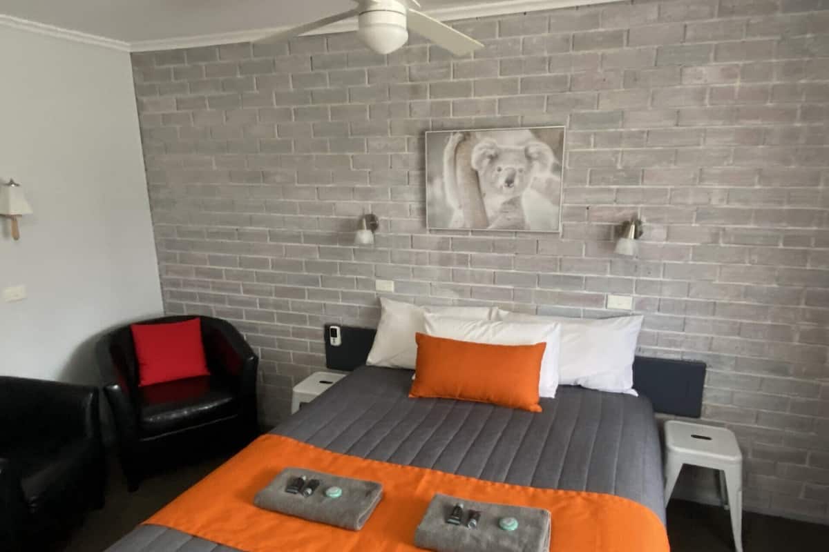 A hotel room with a brick wall, a bed with an orange blanket, a black leather chair, and a ceiling fan.