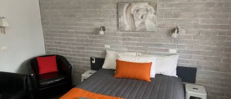 Una habitación de hotel con pared de ladrillo, una cama con manta naranja, una butaca de cuero negro y un ventilador de techo.