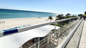 A beach view from a balcony with a clear sky, palm trees, and a sandy beach.