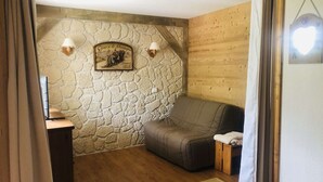 A cozy room with a stone wall, wooden paneling, a sofa, and a small table with a lamp. - La Clairière 1 - Cosy studio with a beautiful view of the resort (LES AVANCHERS VALMOREL)