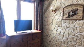 A room with a wooden dresser, a television on top, a stone wall with a family portrait, and a window with curtains. - La Clairière 1 - Cosy studio with a beautiful view of the resort (LES AVANCHERS VALMOREL)