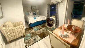Un dormitorio con cama, una silla, una mesa con una vela y una ventana con cortinas.