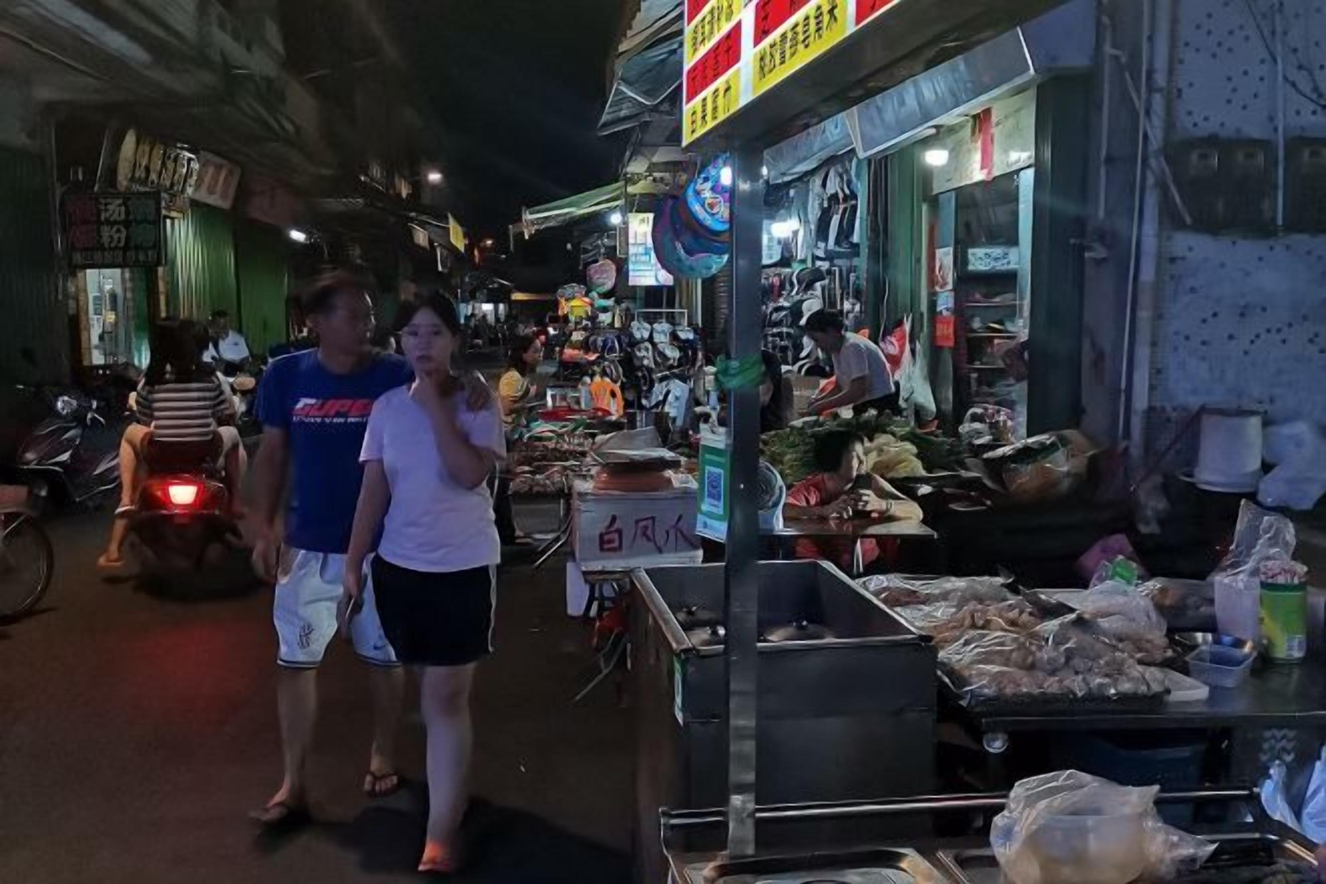 A nighttime street scene with pedestrians, a market stall with food items, and a sign with Chinese characters.