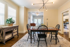 A dining area with a wooden table, four chairs, a chandelier, and a kitchen visible through an open door.