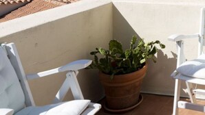 Un balcón con una silla blanca acolchada, una silla de madera, una mesa con mantel blanco y una planta en maceta.
