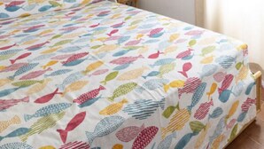 Una cama con una colcha de estampado de peces de colores, una mesita de noche con lámpara y una ventana con cortinas.