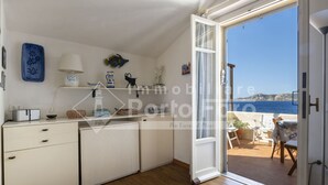 Una cocina compacta con armarios blancos, una pequeña encimera y vista al mar desde un ventanal abierto.