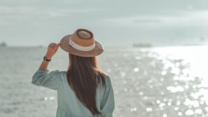 Uma pessoa de vestido azul claro à beira-mar, segurando um chapéu de palha.