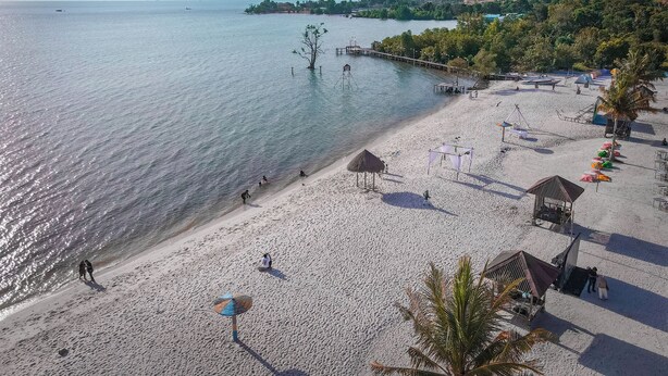 Uma praia com um píer que se estende por águas calmas, rodeada por vegetação e algumas cabanas de praia.
