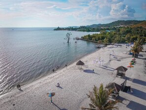 Una playa con un muelle que se adentra en aguas tranquilas, rodeada de vegetación y algunas cabañas de playa.