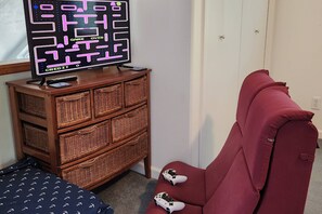 A room with a television displaying a maze game, a wooden dresser with baskets, a red chair, and a bed with a patterned bedspread. - Neskowin Beach Escape, Family & Pet Friendly, Fully Fenced Backyard, EV Charger, Short Walk to Beach (Neskowin)
