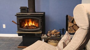 Een kamer met een zwart houtkacheltje, een stoel en een stapelhout tegen een blauwe muur.