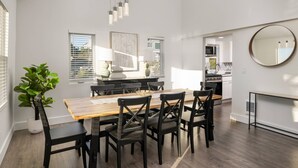 Uma área de jantar moderna com uma mesa de madeira e cadeiras pretas, uma janela grande com persianas e uma cozinha visível ao fundo.