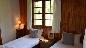 A room with two single beds, wooden walls, a window with curtains, a bedside lamp, and a small table with a picture frame and a book.
