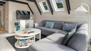 Una acogedora sala de estar tipo loft con un sofá gris, una mesa de centro blanca con libros, un televisor de pantalla plana y una lámpara de pie blanca.