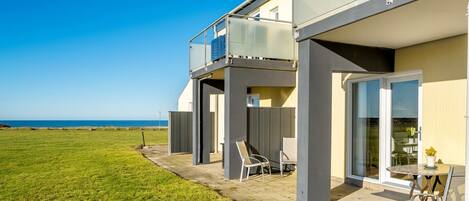 A modern two-story house with a balcony overlooking a grassy area and the sea.