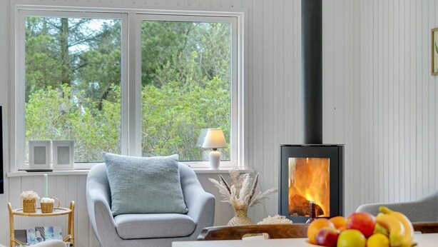 Ein gemütliches Zimmer mit großem Fenster, einem modernen Kamin, einem bequemen Sessel und einer Obstschale auf dem Tisch.