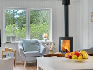 Een knus ingerichte kamer met een groot raam, een moderne haard, een comfortabele fauteuil en een schaal fruit op de tafel.