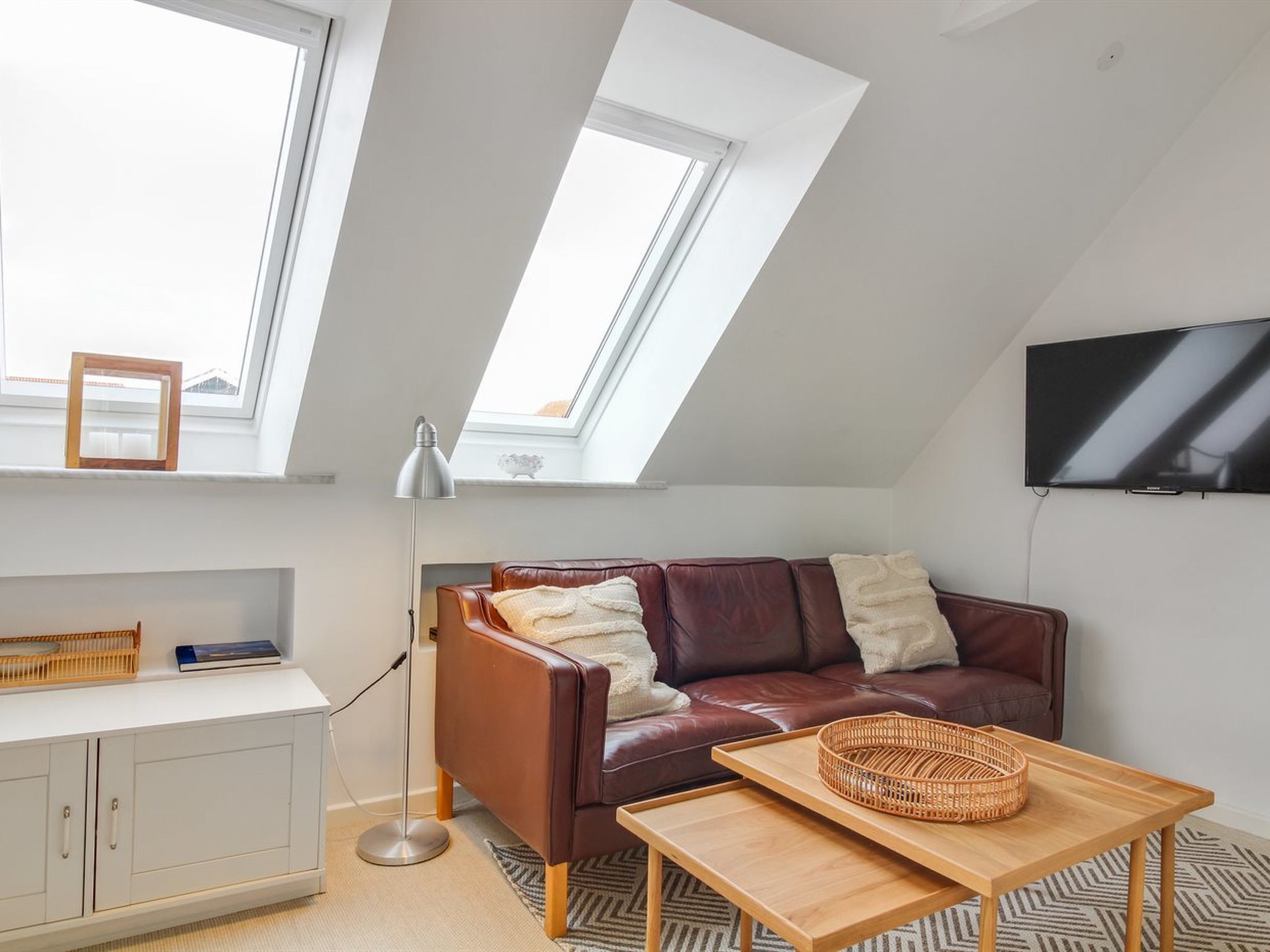 A modern loft apartment with a sloped ceiling, a brown leather sofa, a wooden coffee table, and a flat-screen TV mounted on the wall.