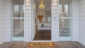 A modern entrance with a 'Welcome' mat, a chandelier, and a view into a room with a wooden floor and a white cabinet.