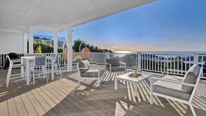 A covered outdoor deck with white furniture, a grill, and a view of the ocean.