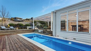 A modern outdoor pool area with a wooden deck, a covered seating area, and a large windowed room.