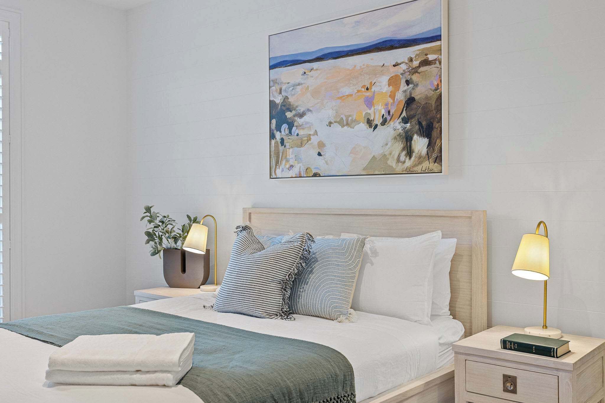 A neatly made bed with pillows, a bedside lamp, a book, and a painting on the wall.