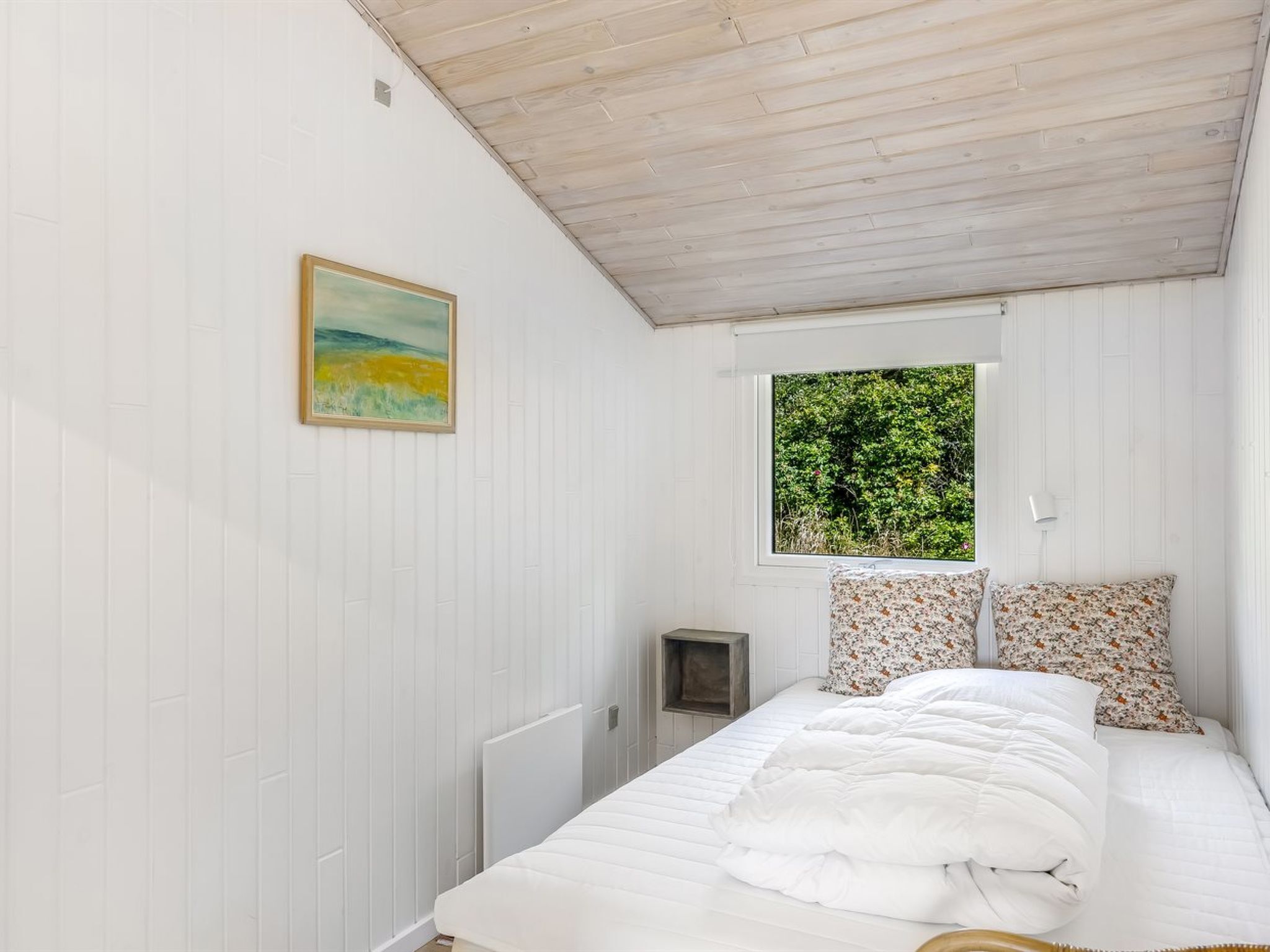 Ein Schlafzimmer mit einem Bett, einem Gemälde, einem Fenster mit Blick auf Grünflächen und einem kleinen Nachttisch.