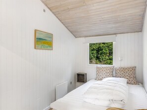 A bedroom with a bed, a painting, a window with a view of greenery, and a small bedside table.