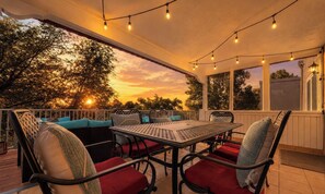 Eine Terrasse im Freien mit Tisch, Stühlen, Lichterketten und Blick auf Bäume und Sonnenuntergang.