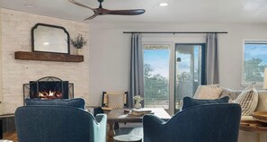 Ein Wohnzimmer mit Kamin, Deckenventilator, Balkon mit Vorhängen und einer Couch mit Kissen.