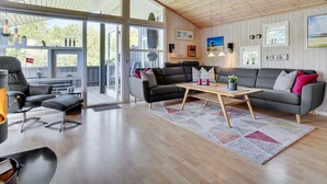 Amplia sala de estar con un sofá grande, una mesa de centro y una chimenea. Varias ventanas ofrecen luz natural y vistas al exterior.
