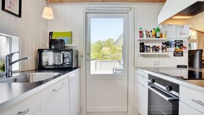 Cocina compacta con armarios blancos, microondas negro, pileta de acero inoxidable y horno negro. Una ventana ofrece vista a la vegetación exterior.