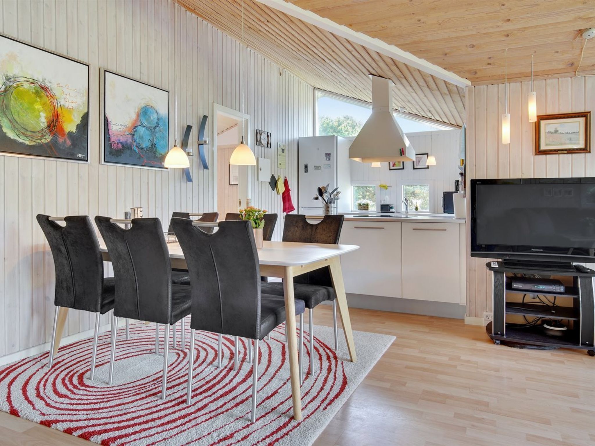 Een moderne keuken met witte kasten, een eethoek met een witte tafel en zwarte stoelen, en een grote flatscreen televisie.