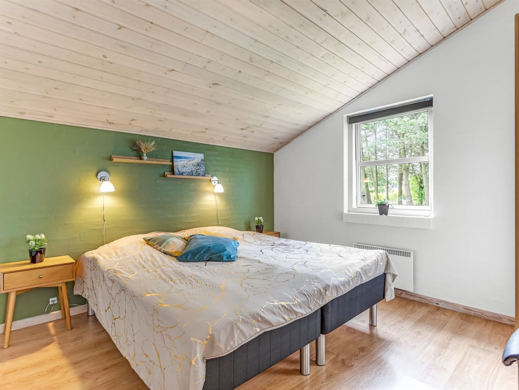 A bedroom with a sloped ceiling, a large bed with a grey upholstered headboard, a window with a view of trees, and a small bedside table with a lamp.