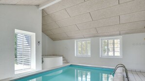 A swimming pool with clear blue water, a tiled floor, and a ceiling with square tiles. There are windows on the walls, and a staircase leading to an upper level.