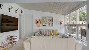 A living room with a white sofa, a flat-screen TV, a wall-mounted shelf with plates, and a window with a view of the outdoors.