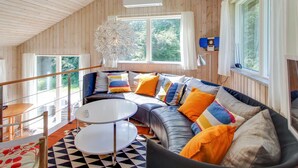 A cozy cabin interior with a wooden ceiling, a large window, a sofa with colorful cushions, and a round table.