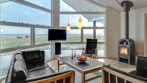 Een moderne woonkamer met een groot raam dat uitkijkt op een strand, een open haard en een eethoek met een schaal fruit.