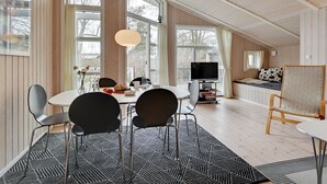 Een ruime kamer met een hoge plafond, grote ramen, een eethoek met een tafel en stoelen, een televisie en een gezellige zithoek met een bank en fauteuil.