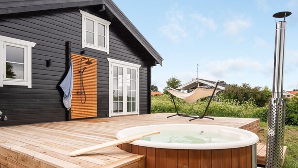 Un terrazzo in legno con una vasca idromassaggio e una sauna, una casa con facciata scura e finestre bianche, e un'amaca sullo sfondo.