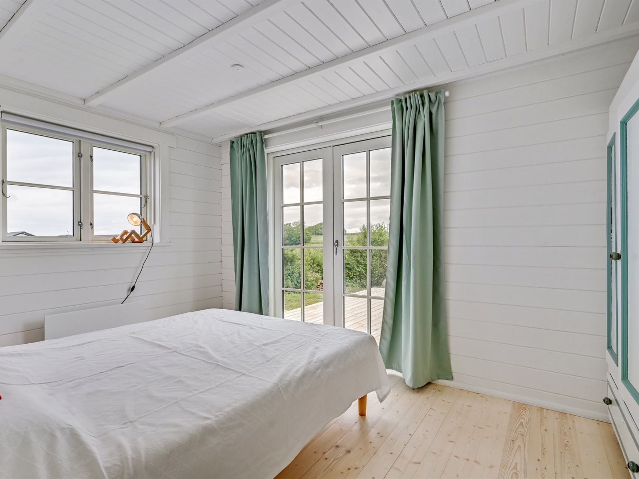 Ein Schlafzimmer mit Bett, Fenster und Blick auf Grünfläche.