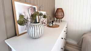 Une pièce bien rangée, avec une commode blanche, un tableau encadré, un vase blanc contenant des plantes vertes, une bougie et une lampe en osier.