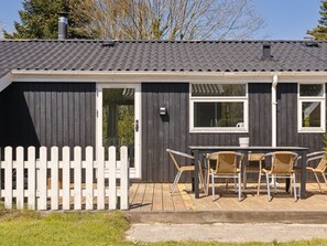A small, dark wooden house with a tiled roof, a white picket fence, and a patio with wicker chairs and a table.