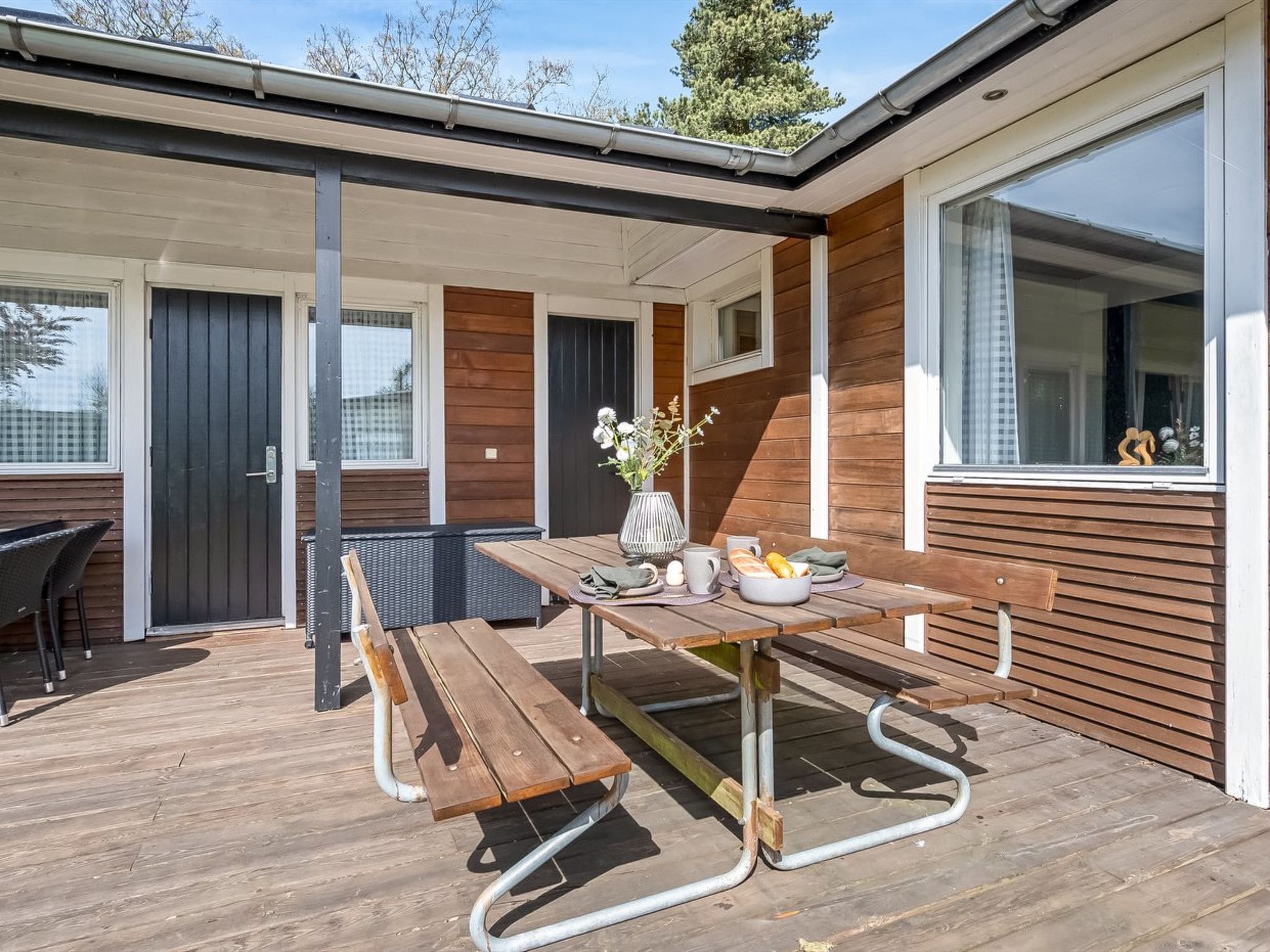 A wooden deck with a picnic table and benches, a potted plant, and a window with a view of trees.