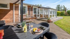 A wooden deck with a table set for dining, a glass-enclosed patio area, and a view of the surrounding greenery. - "Teija" - 300m from the sea by Interhome (Odder)