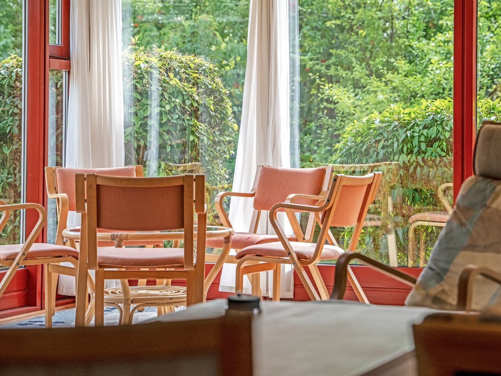 A room with a view of greenery through large windows, wooden chairs, and a table.