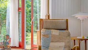 Ein Zimmer mit Holzstuhl, einem kleinen Tisch und einer Lampe. Ein Fenster mit Vorhängen bietet Blick auf grüne Landschaft.